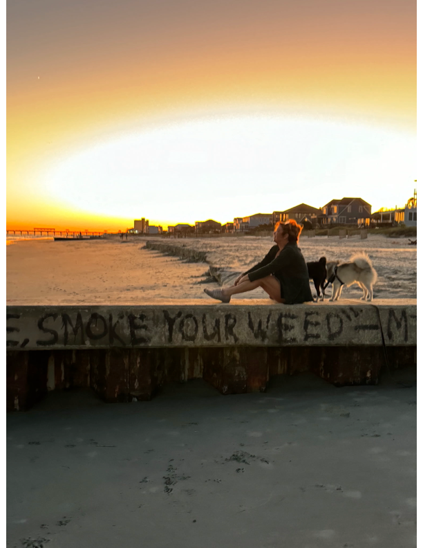 Doreen sitting on beach with sunset behind, and "Smoke Your Weed" sign below